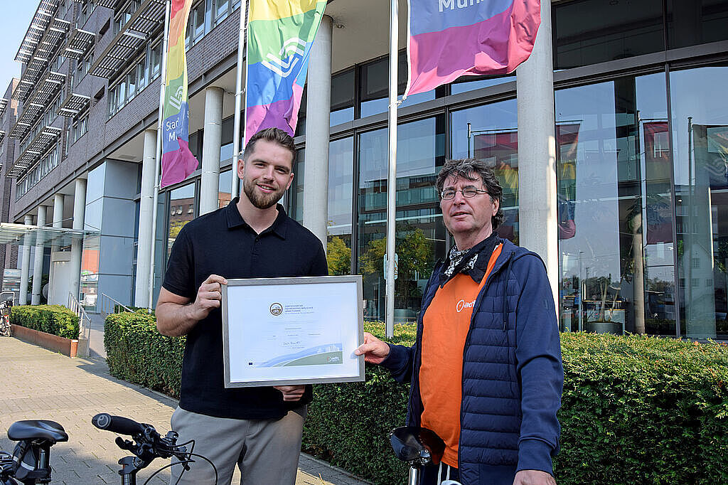2024_09_18_ffa_Stadtwerke Muenster_Florian Adler Zwei Männer stehen vor einem Bürogebäude. Der rechts stehende Mann reicht dem links stehenden Mann ein eingerahmtes Zertifikat. Im Hintergrund wehen Fahnen in Regenbogenfarben. Im Vordergrund stehen zwei Fahrräder.