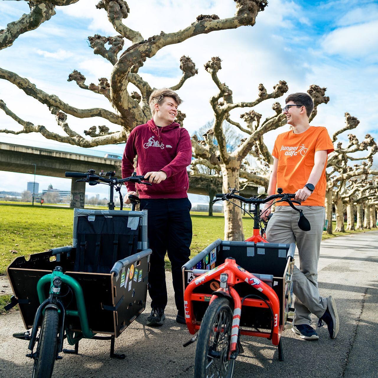 Cargobike Duo Landesjugendversammlung 2026 Zwei junge Männer stehen mit Cargobikes auf einem Gehweg, im Hintergrund ist eine Baum-Allee und eine Brücke.
