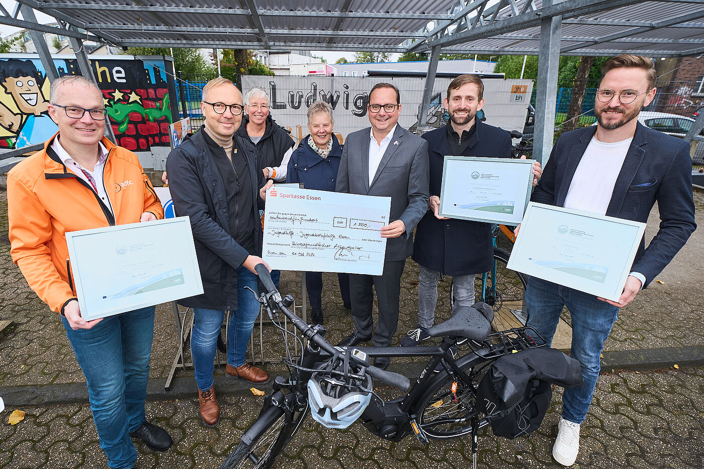 2024_10_01_FFA_Jugendhilfe Essen_Moritz Leick, Stadt Essen Auf dem Bild sind sieben Personen zu sehen. Einige von Ihnen halten gerahmte Urkunden in die Kamera. Im Vordergrund des Bildes steht ein Fahrrad.