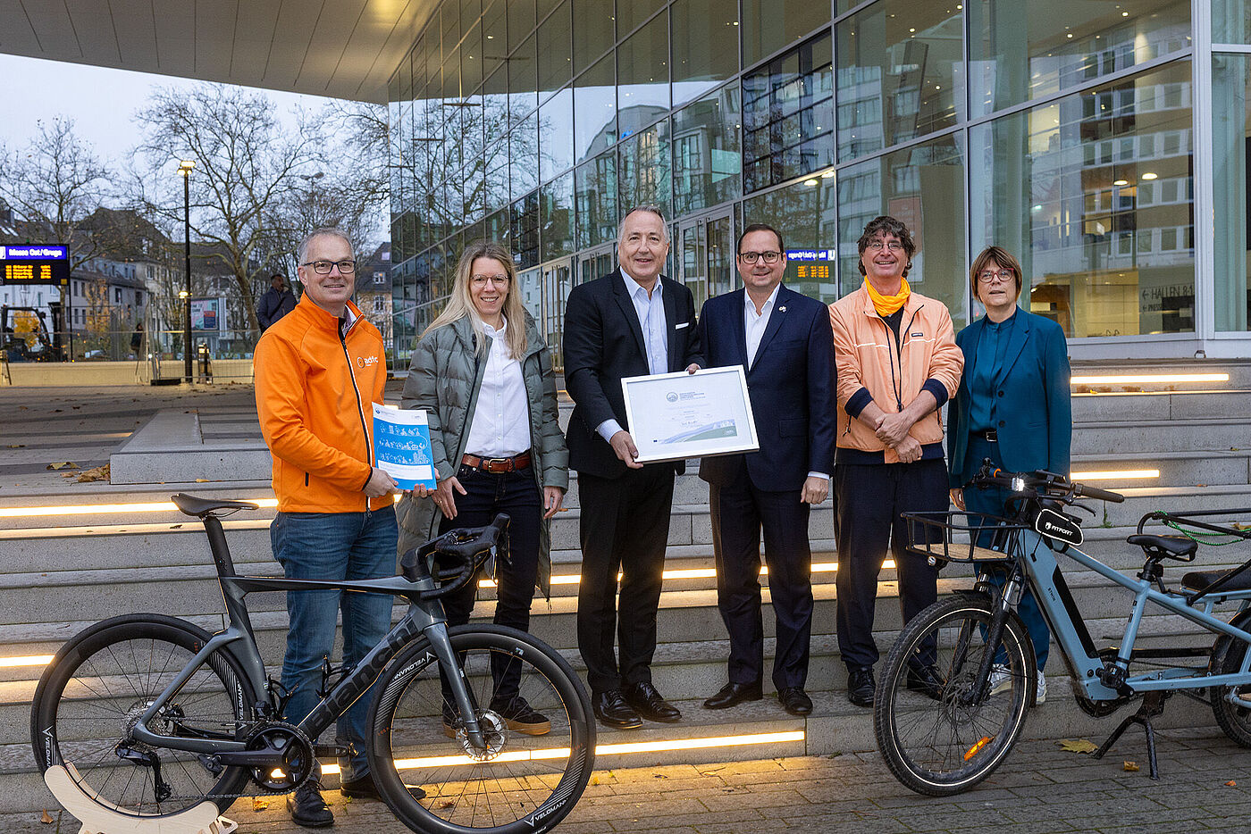 2024_11_26_FFA_Messe Essen_C_Georg Lukas Sechs Personen stehen vor einem Gebäude mit Glasfassade. In der Mitte stehen zwei Männer, die gemeinsam ein gerahmtes Zertifikat hochhalten. Im Vordergrund stehen zwei Fahrräder.