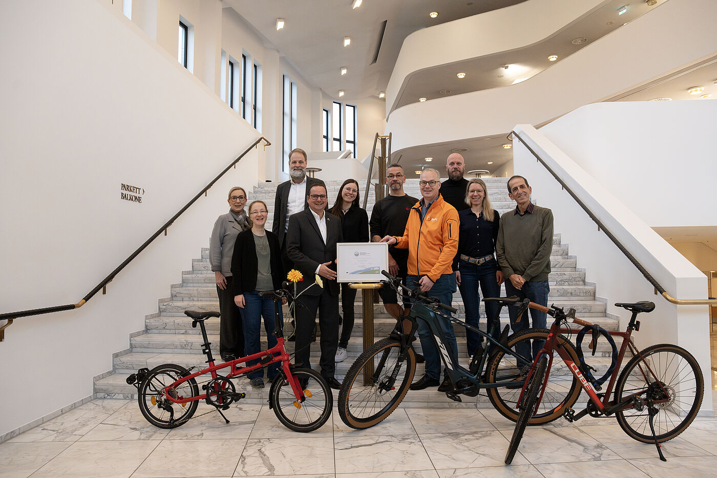 2024_11_11_FFA_TUP Essen_C_Caroline Schlüter Zehn Menschen stehen auf einer weißen Treppe. Ein Mann in Anzug und ein Mann in einer orangenen Jacke halten ein gerahmtes Zertifikat. Die anderen Menschen stehen um die beiden Männer herum. Im Vordergrund sind Fahrräder zu sehen. Im Hintergrund sieht man weitere Treppen und Ballustraden.
