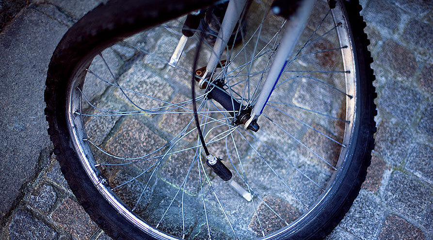 Nahaufnahme eines Fahrradvorderrads auf Kopfsteinpflaster. Die Felge ist verbogen und mehrere Speichen sind gebrochen oder hängen lose herab. Das Rad steht schief auf dem grauen Pflaster und ist offensichtlich nach einem Unfall nicht mehr fahrbereit.